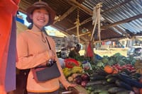 A traveler posing at a bustling open-air market stall stocked with various fresh fruits and vegetables.