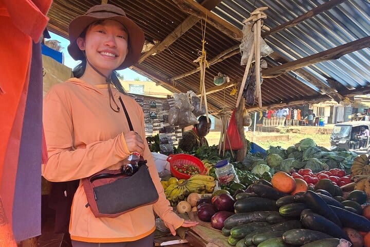 A traveler posing at a bustling open-air market stall stocked with various fresh fruits and vegetables.