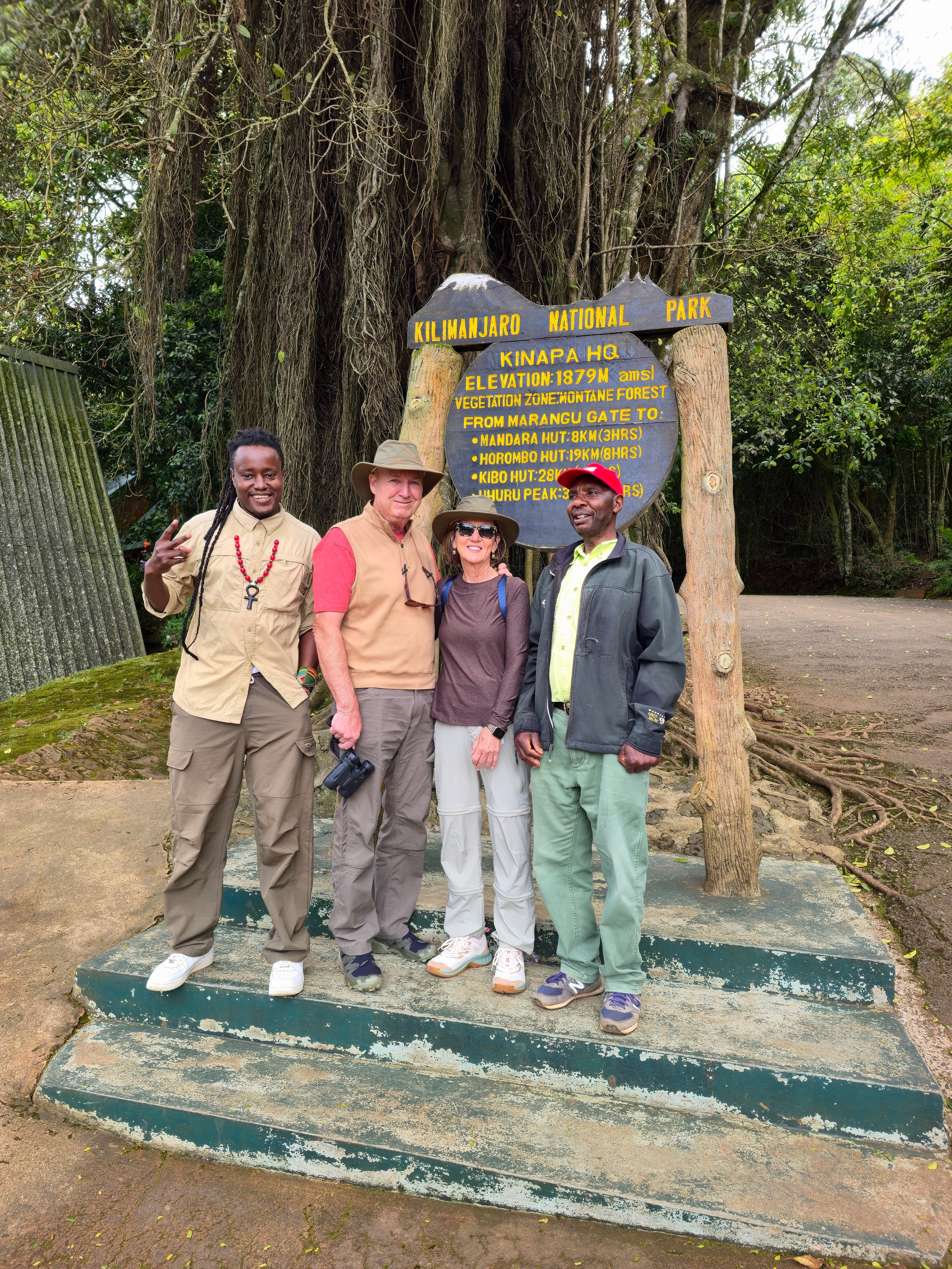 A group of four travelers and local guides standing together for a photo next to the wooden park entrance sign on Mount Kilimanjaro.