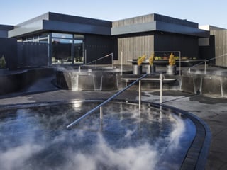 Exterior view of Krauma Geothermal Baths at dawn or dusk, showing multiple steaming circular pools set in dark stone terracing, with the ang