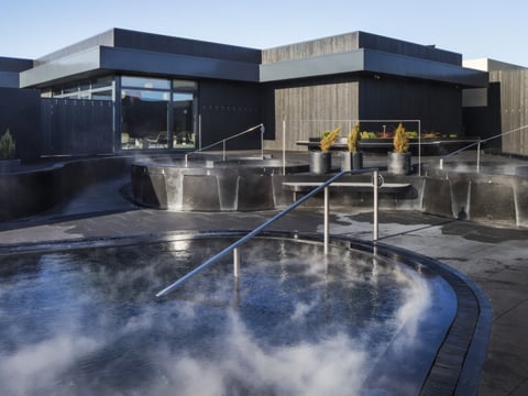 Exterior view of Krauma Geothermal Baths at dawn or dusk, showing multiple steaming circular pools set in dark stone terracing, with the ang