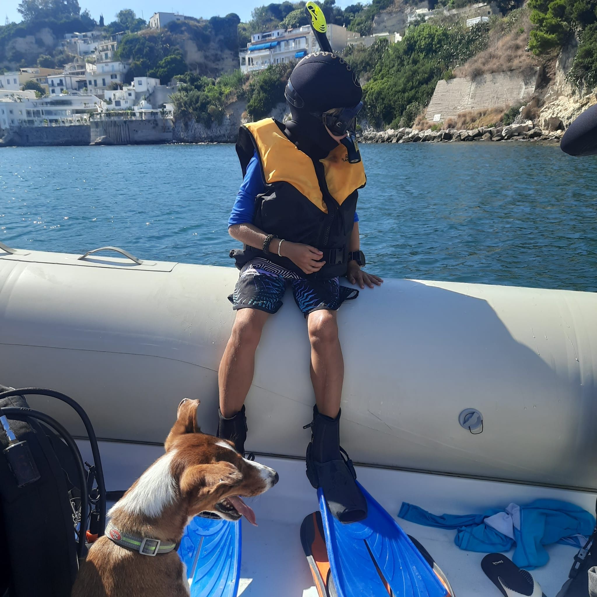 A young kid all dressed up in snorkeling equipment, with a life jacket, getting ready to go in the water from the boat, next to his dog..