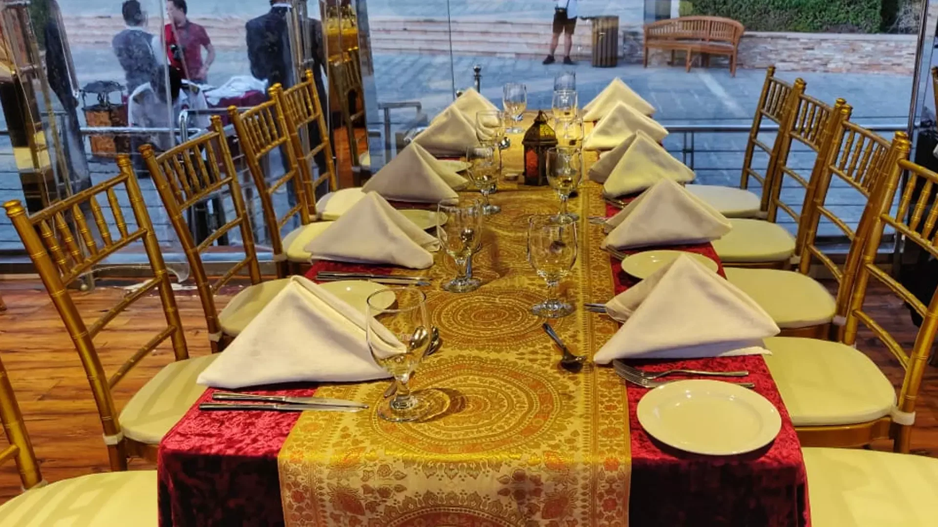 A long dining table is set with folded white napkins, glassware, and gold chairs inside a venue overlooking an outdoor walkway at Dubai Water Canal Dhow Cruise.