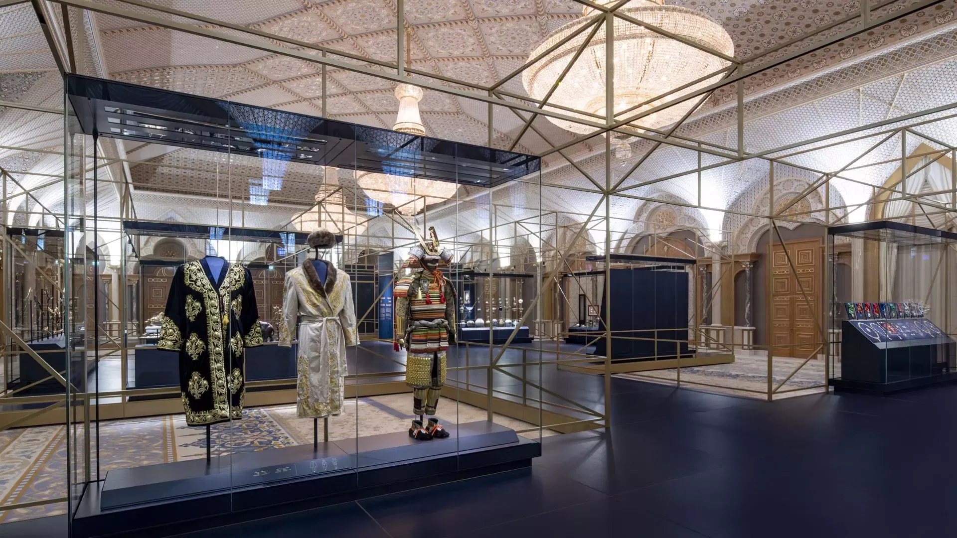 A glass display case in a grand, ornate hall holds a collection of traditional garments including an embroidered black robe, a white robe, and a suit of armor at Qasr Al Watan, Royal Palace.