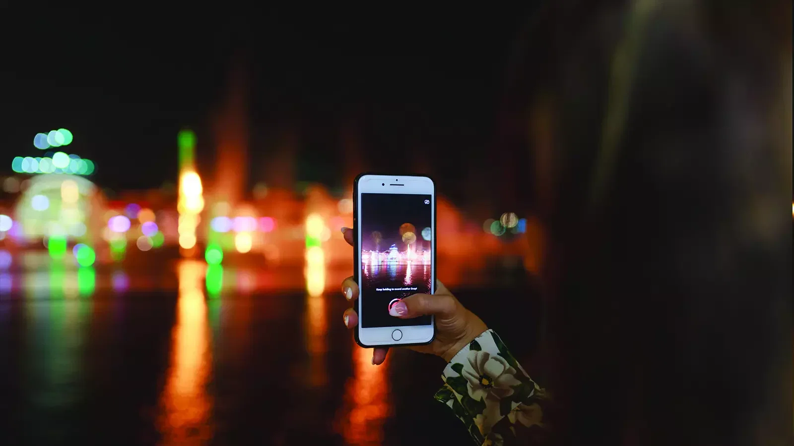 A person holds up a smartphone to record a nighttime cityscape with colorful blurred lights reflecting on water at Dubai Water Canal Dhow Cruise.