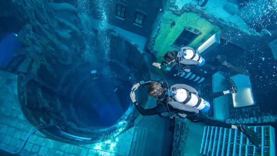 Two scuba divers swim past a sunken urban structure with a large circular opening and a tiled floor at Deep Dive Dubai.