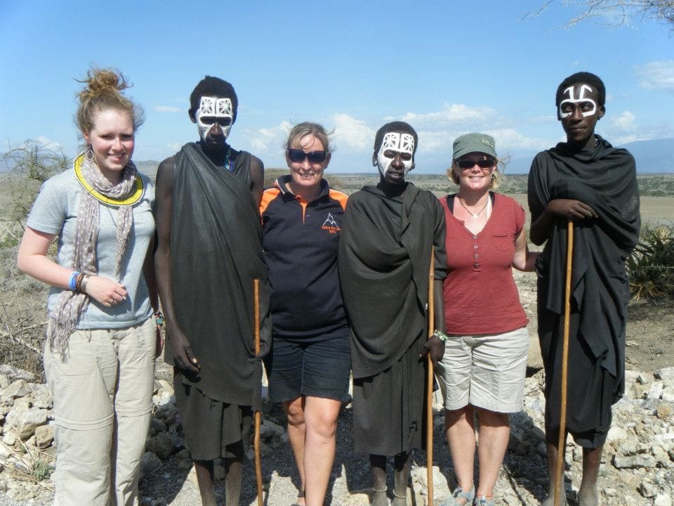 A group of female tourists standing alongside Maasai initiates dressed in traditional dark robes with white face paint patterns outdoors.