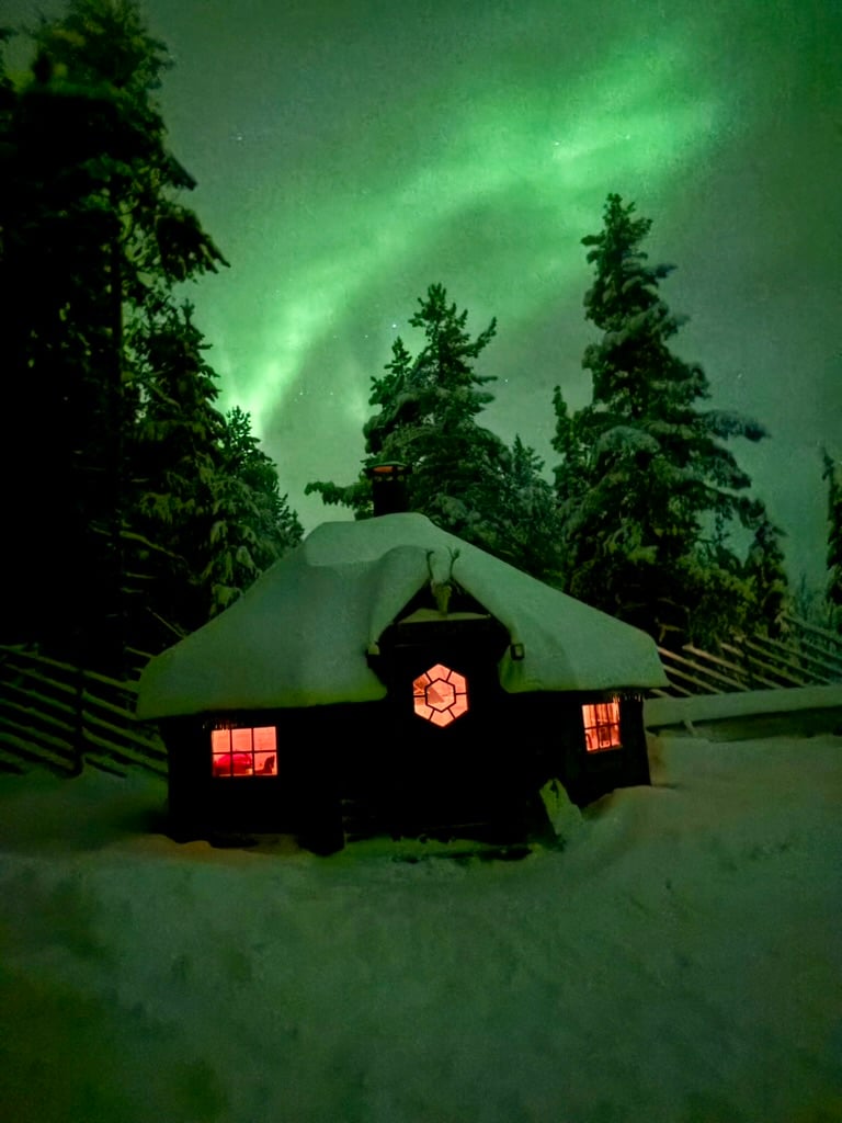 A warmly illuminated lakeside kota surrounded by deep snow beneath the Northern Lights.