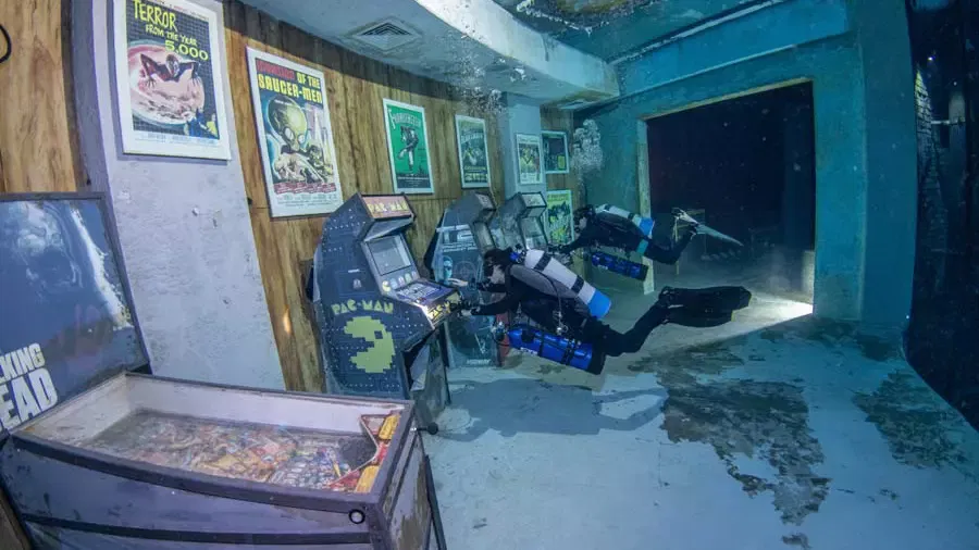 A scuba diver floats in front of vintage arcade cabinets and movie posters mounted on a submerged wooden wall at Deep Dive Dubai.
