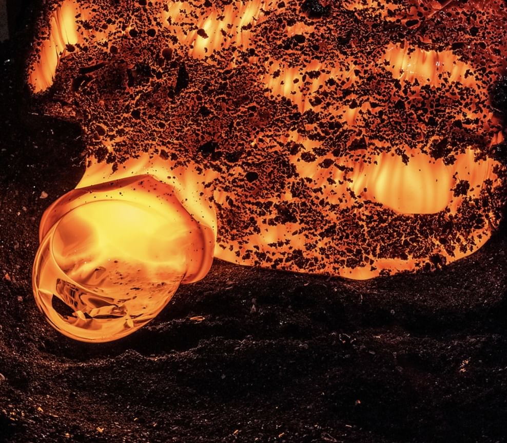 Close-up of a glowing molten lava formation bubbling on a black sandy surface, captured in vivid orange and red tones.