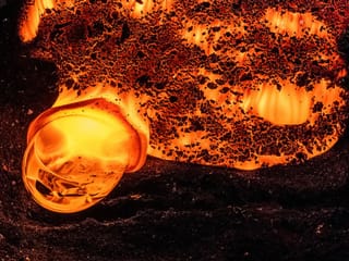 Close-up of a glowing molten lava formation bubbling on a black sandy surface, captured in vivid orange and red tones.