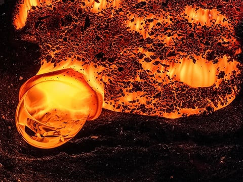 Close-up of a glowing molten lava formation bubbling on a black sandy surface, captured in vivid orange and red tones.