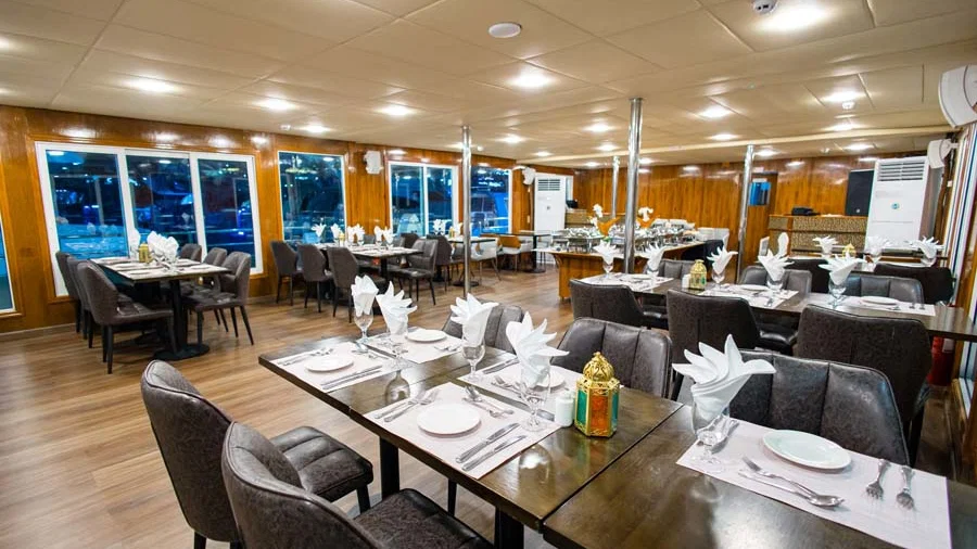A wide-angle view of the wood-paneled dining room shows neatly set tables with folded napkins and golden decorative lanterns, surrounded by large glass windows at Alexandra Sea Lounge.