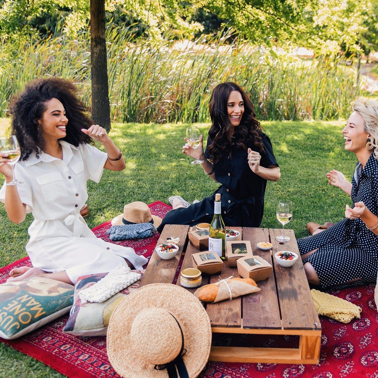 Group of friends sitting on picnic blankets under oak trees at Rhebokskloof Wine Estate, raising wine glasses.