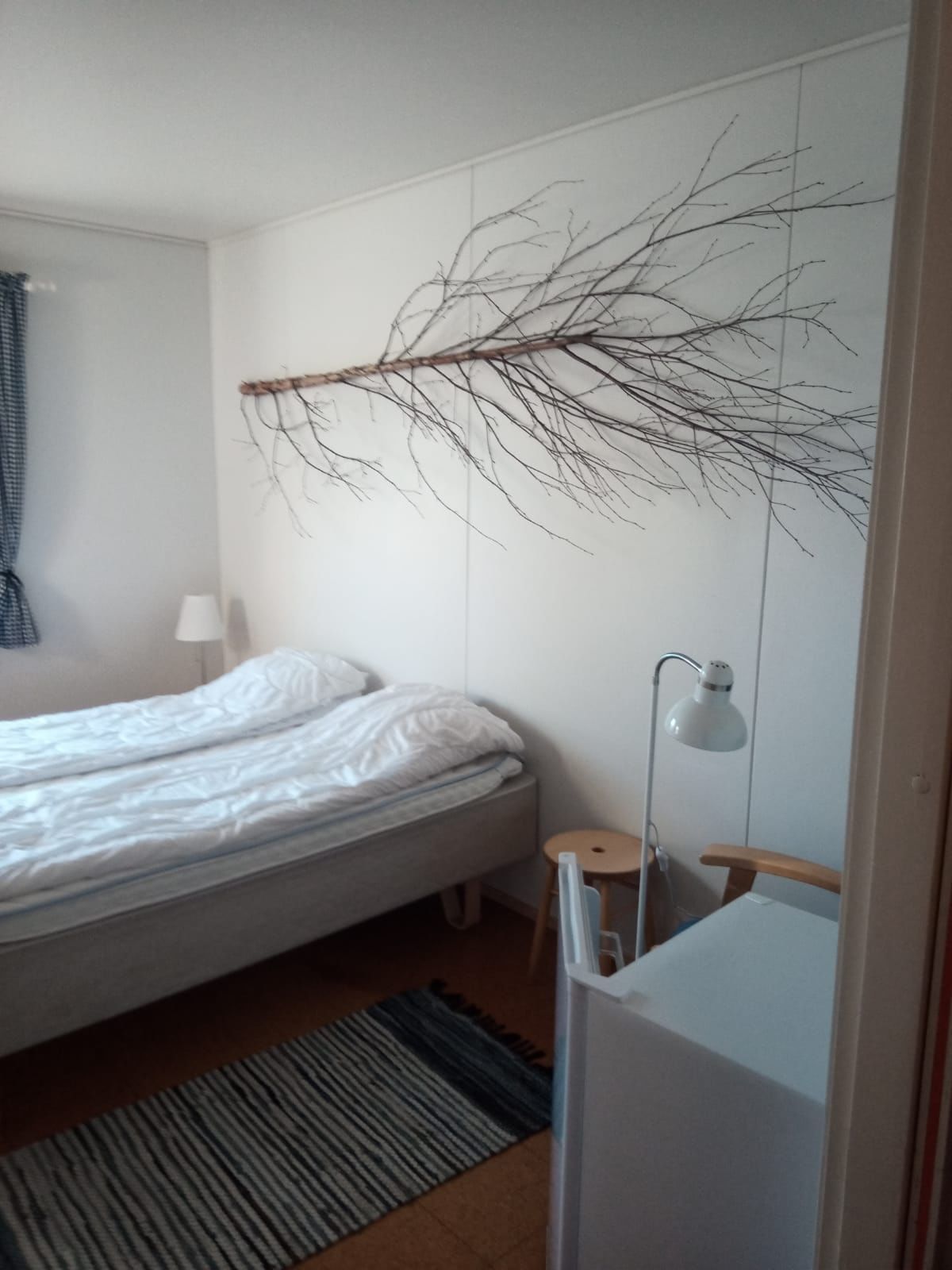 Simple white bedroom with single bed, wooden floor, nightstand, reading lamp, and a decorative branch on the wall