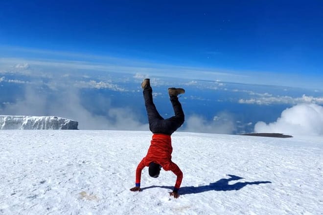 A person in a red jacket performing a handstand on a snowy mountain peak with a blue sky and clouds in the background.