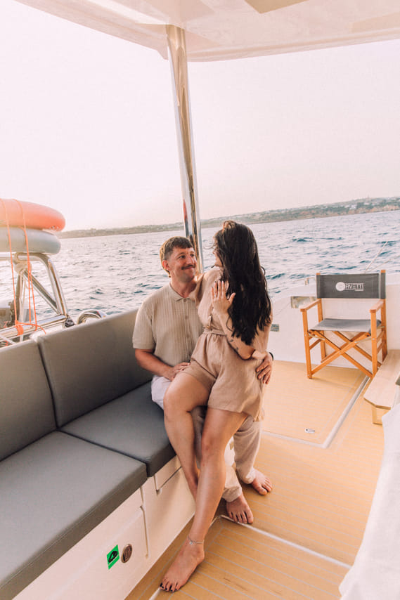 A man and woman sitting close together on a yacht bench, with her sitting on his lap. They are both smiling, positioned behind a set dining 