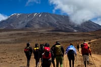 A group of hikers with backpacks walking across a rocky plateau toward a large, snow-capped mountain under a blue sky.