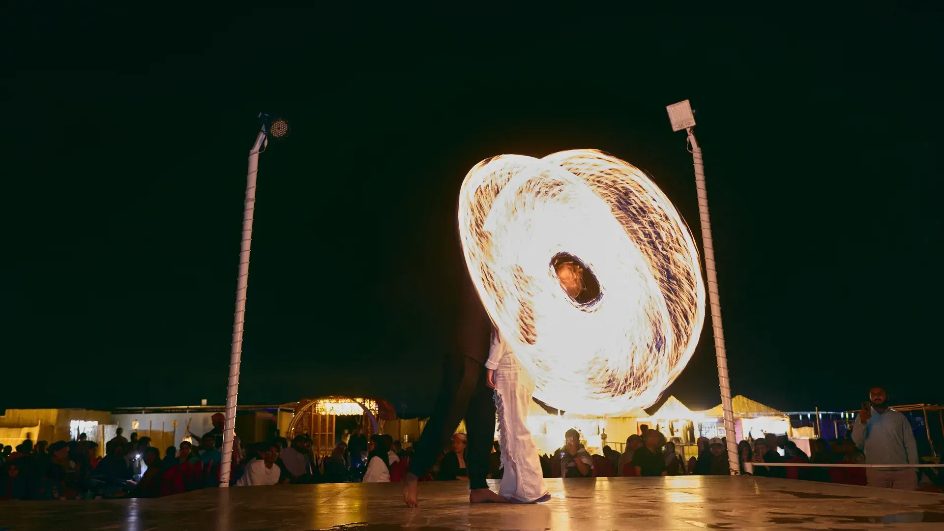 A performer on stage spins a fiery apparatus that creates a glowing circular pattern against the dark background at Desert Safari + Al Faqaa Camp.