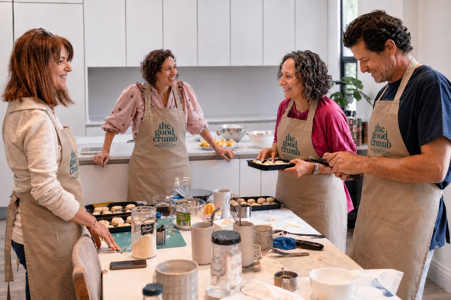 4 people standing around a table, talking, smiling, baking scones