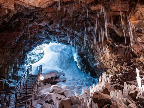 Interior of Vidgelmir lava cave in winter, showing a dramatic arched entrance chamber with ice stalagmites rising from the floor, icicles ha