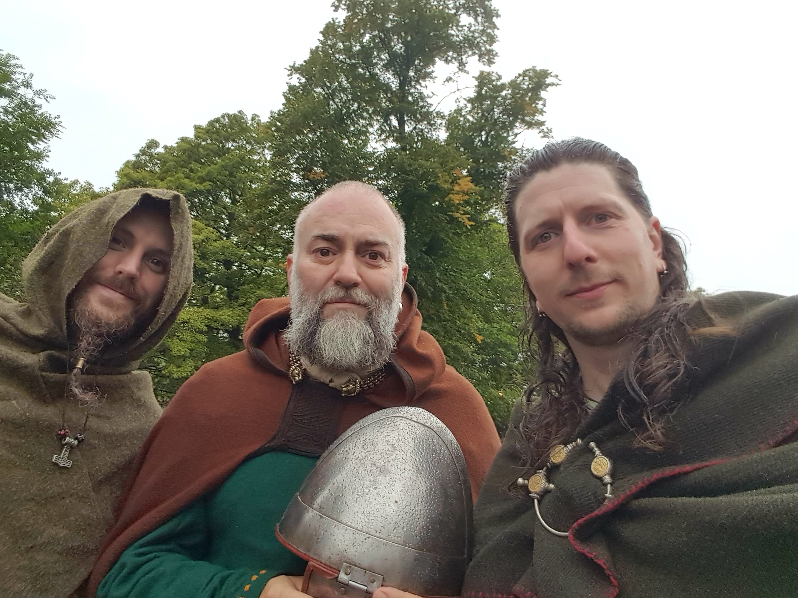 Three viking men stood in the rain, in front of a lush, green tree, getting soggy.