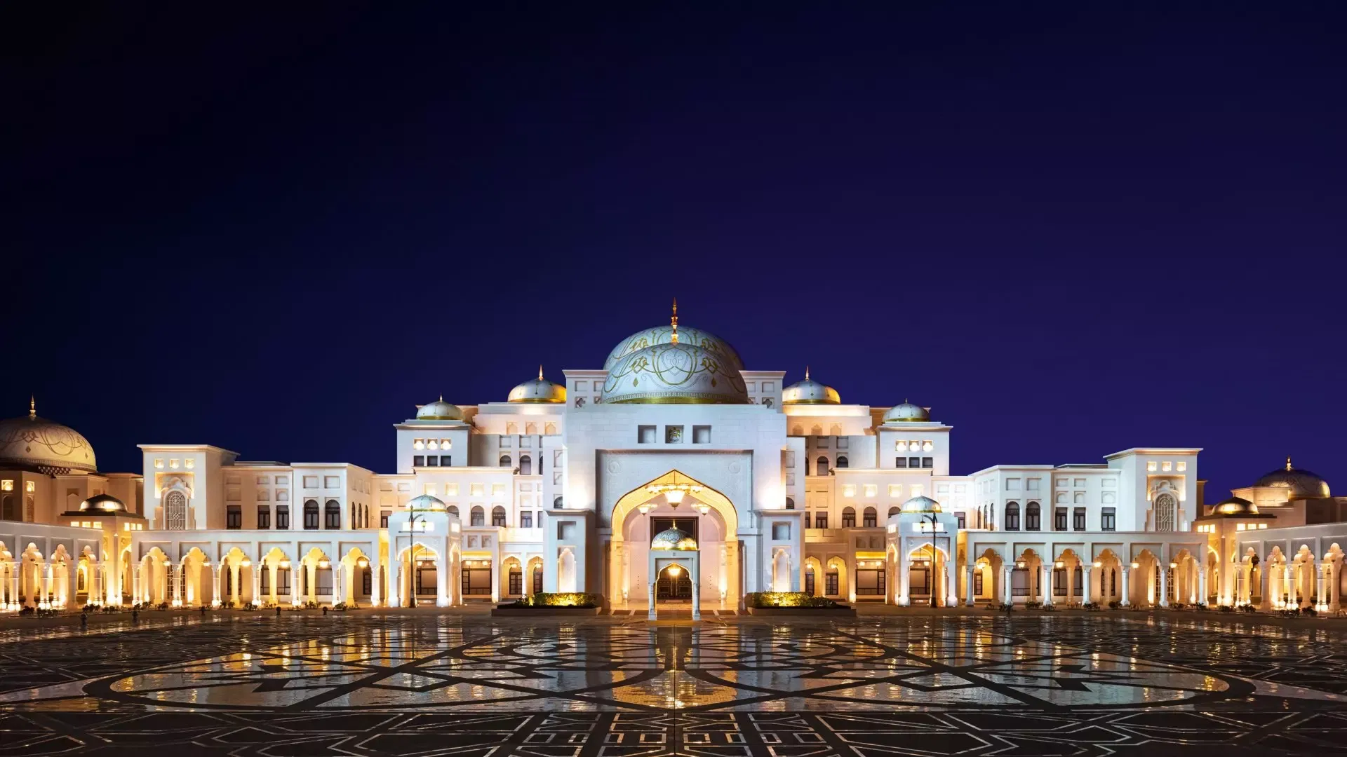 An expansive exterior view of the illuminated palace facade at night, featuring a dark blue sky, golden lighting on the white structure, and reflections on the wet, patterned courtyard floor at Qasr Al Watan, Royal Palace.