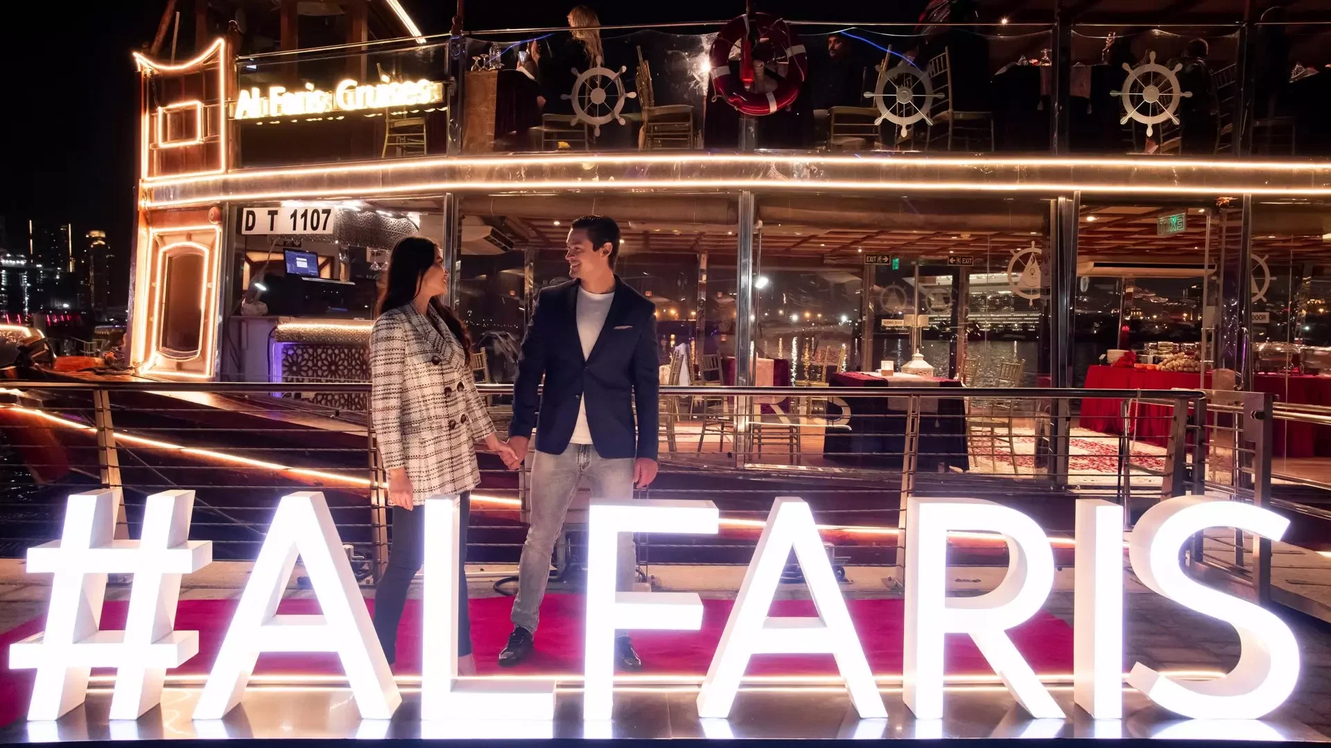 A couple holds hands behind large, illuminated letters on a red carpet in front of a multi-level boat at Dubai Water Canal Dhow Cruise.