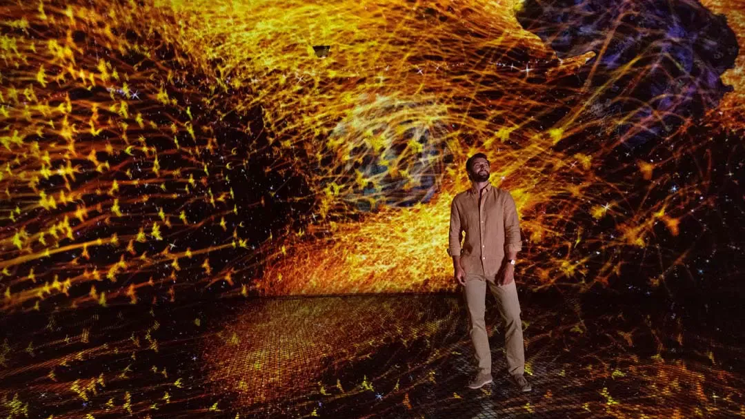 A man stands in an immersive digital room surrounded by glowing golden particle projections at TeamLab Phenomena.