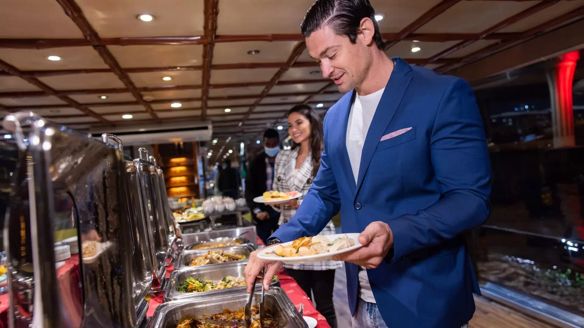 A man in a blue blazer uses tongs to select food from a buffet line on a boat at Dubai Water Canal Dhow Cruise.