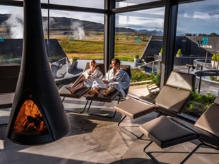 Two guests in white robes reclining on loungers inside Krauma's glass-walled relaxation room, with a suspended wood-burning fireplace in the
