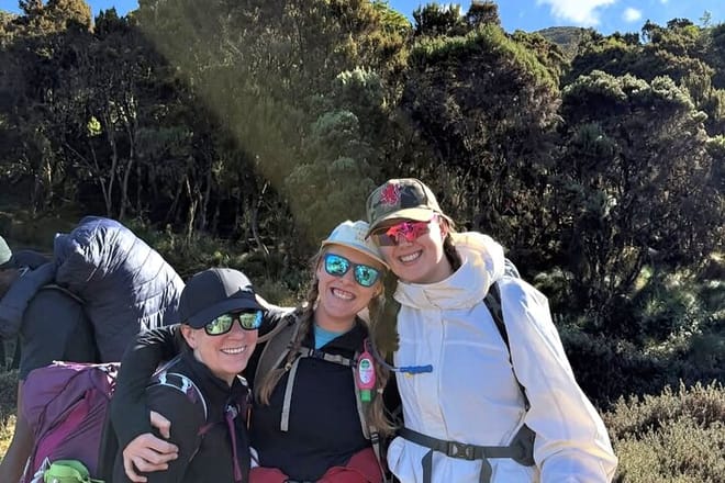 Three smiling hikers wearing sunglasses and hats posing for a photo in front of green mountain vegetation.