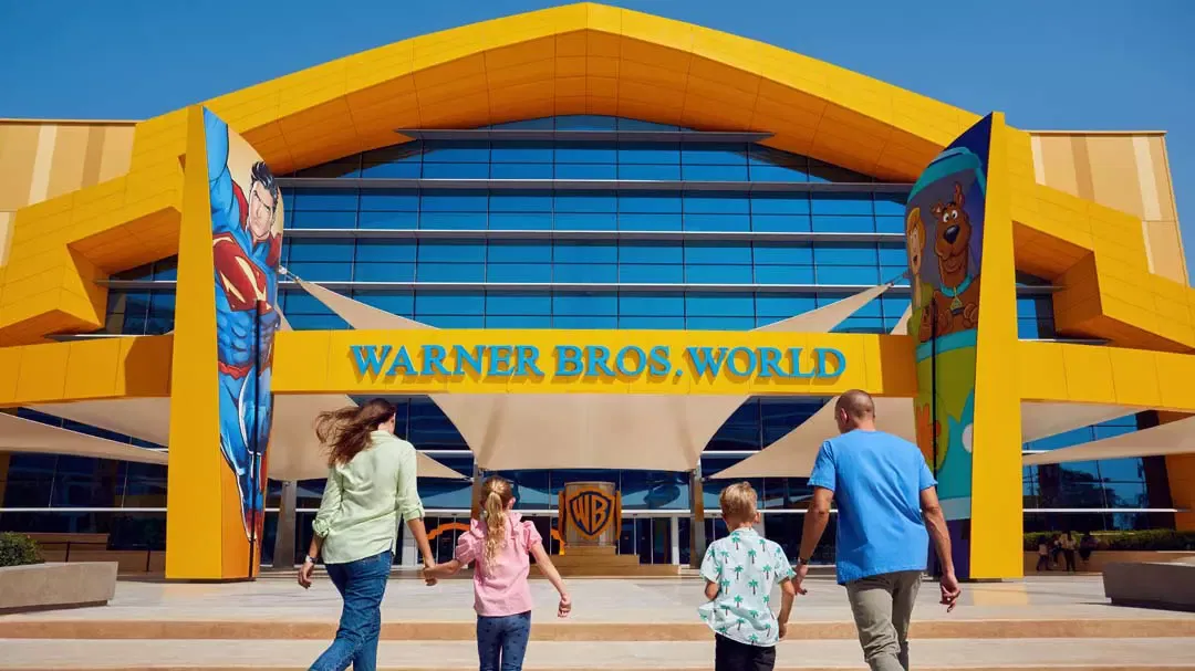 A family of four walks hand-in-hand toward the entrance of a large, yellow-and-blue building at Warner Bros. World Abu Dhabi Theme Park.