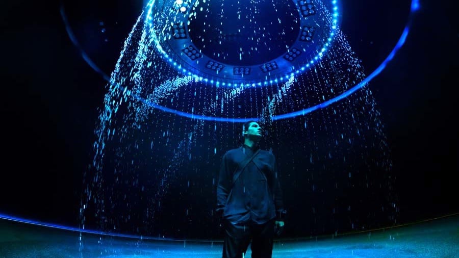 A visitor standing beneath a ring of light as streams of blue light fall around them like rain inside a dark dome at AYA Universe Dubai