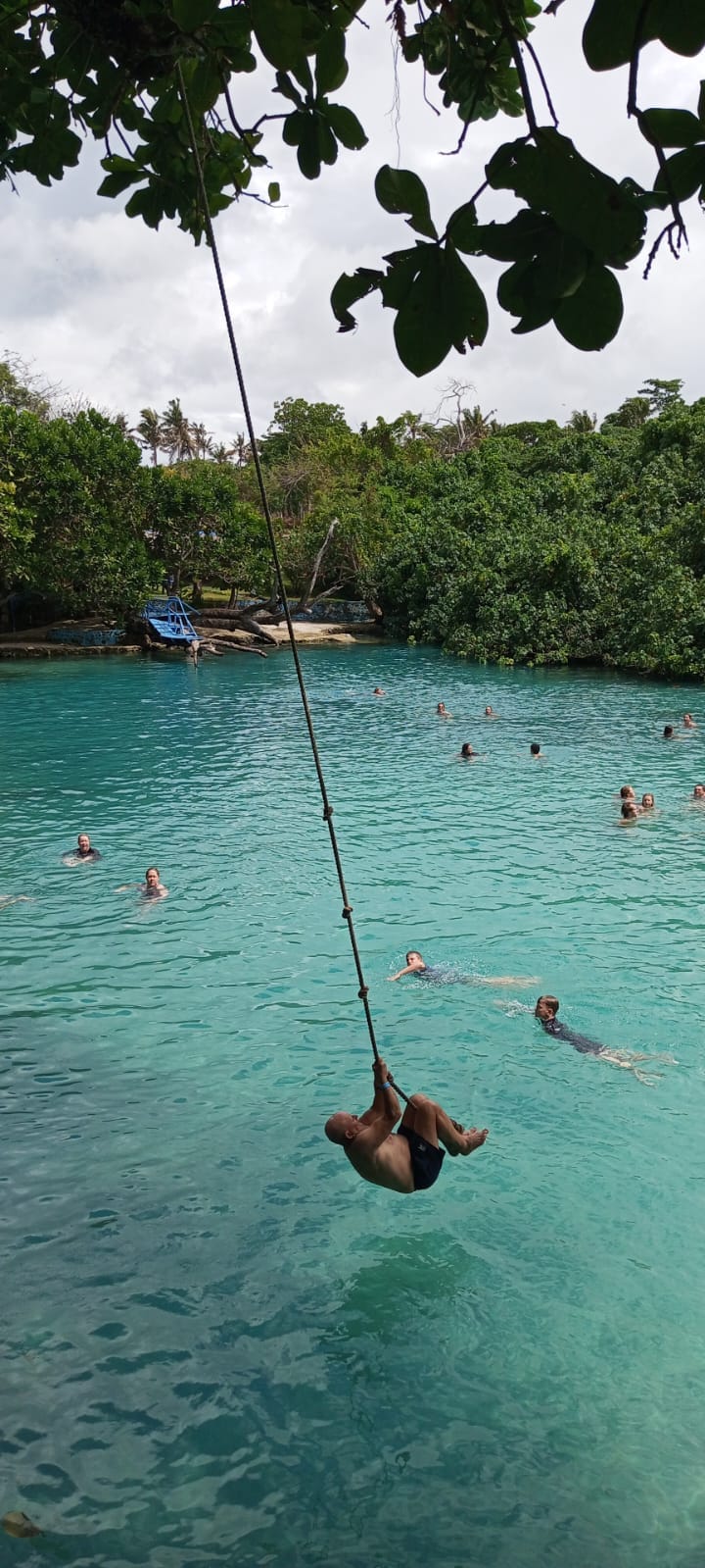 A person swings on a rope above a bright turquoise lagoon while other swimmers float in the water below, surrounded by dense tropical trees.