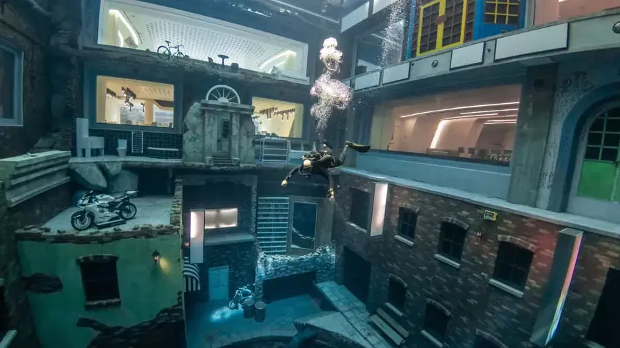 A diver swims past sunken multi-story building facades featuring a submerged motorcycle and windows at Deep Dive Dubai.