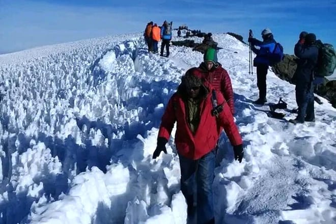 Hikers in winter gear walking along a narrow trail surrounded by jagged white ice formations and snow under a clear blue sky.