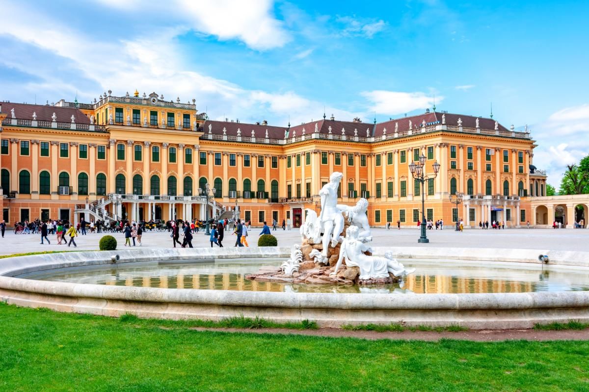 View of Schönbrunn Palace in Vienna with the Neptune Fountain and visitors walking across the courtyard under a blue sky.
