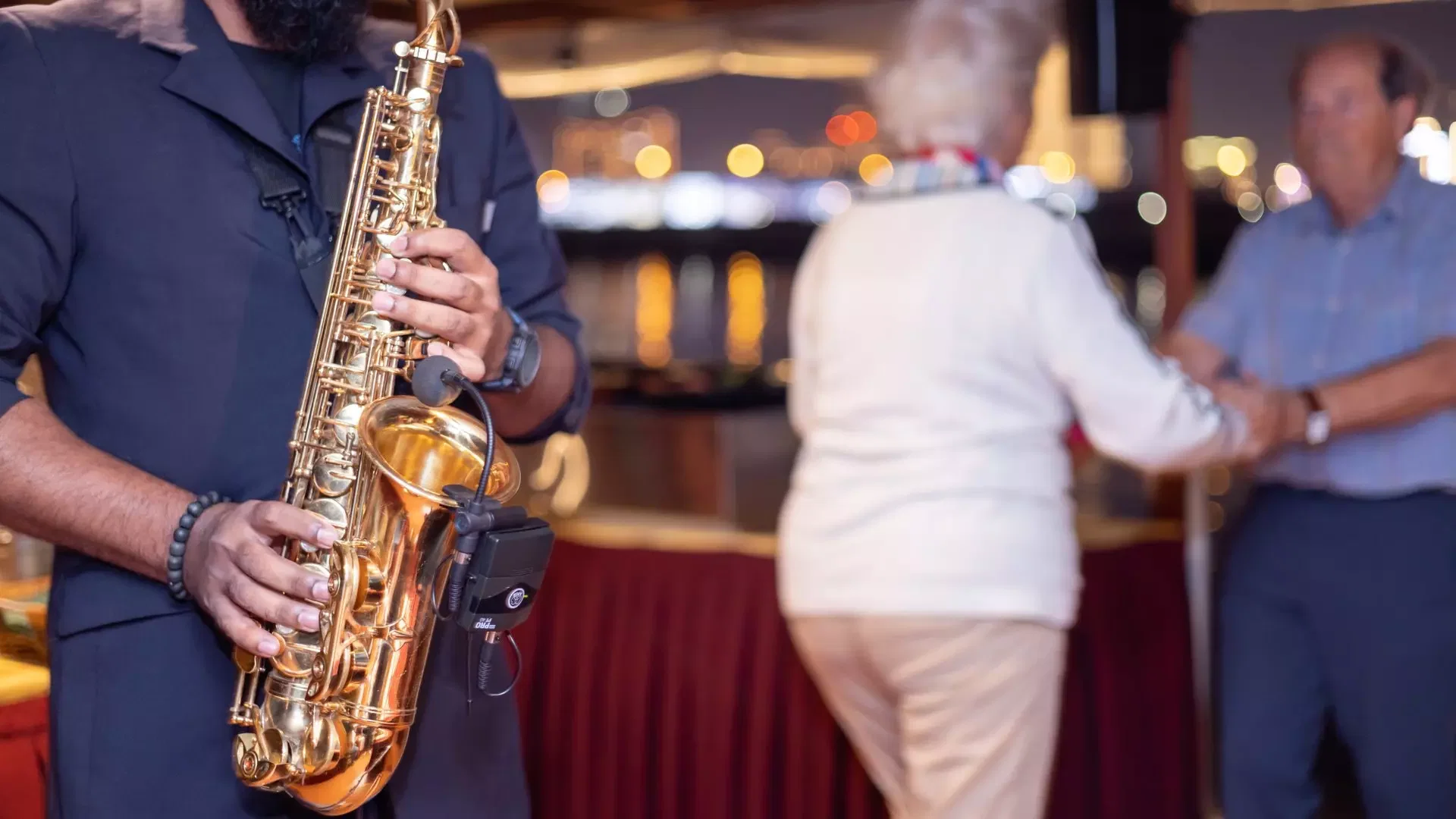 A man plays a gold saxophone while a couple dances in the blurred background at Dubai Water Canal Dhow Cruise.