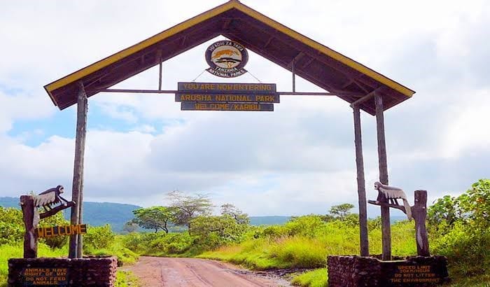A wooden entrance gate sign to Arusha National Park surrounded by green trees and blue sky.