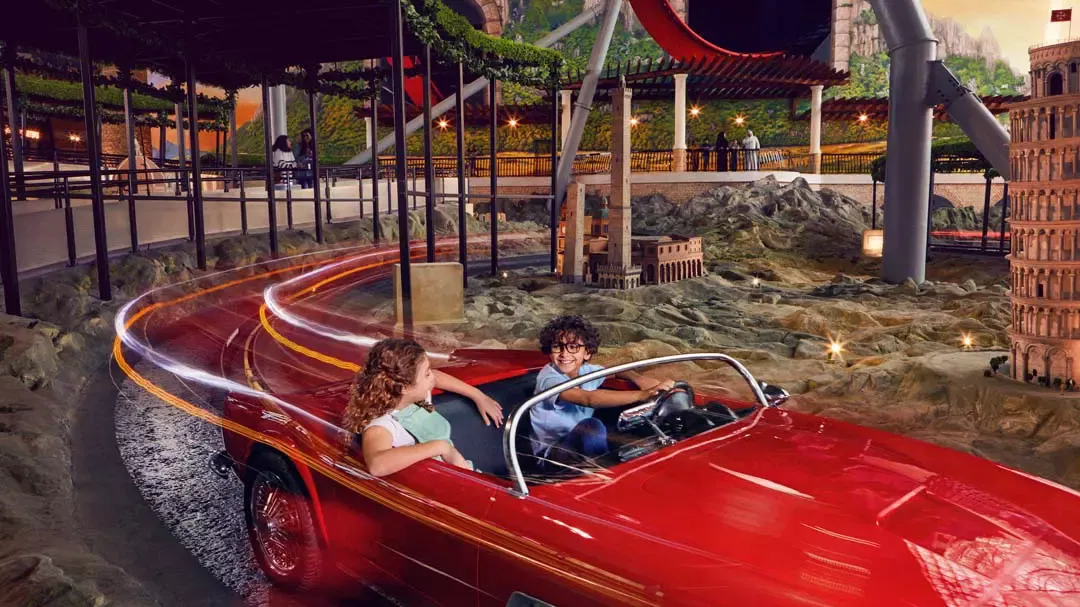 A boy and a girl riding a red classic Ferrari convertible past a miniature Leaning Tower of Pisa in the indoor Italy scene at Ferrari World Abu Dhabi