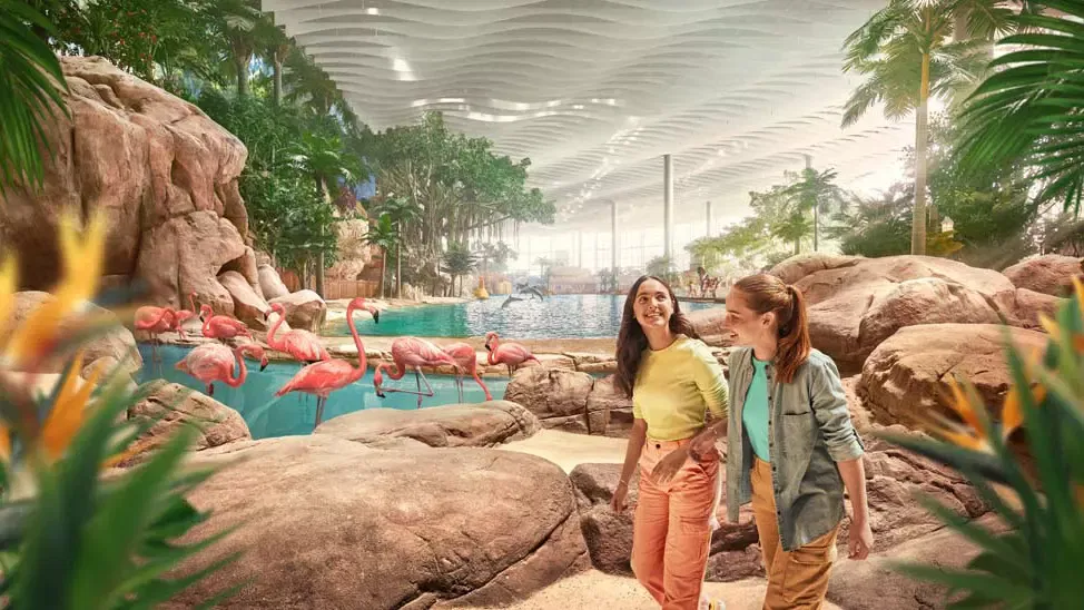 Two young women walk along a rocky path past a pool of pink flamingos in a brightly lit indoor habitat at Sea World Abu Dhabi.
