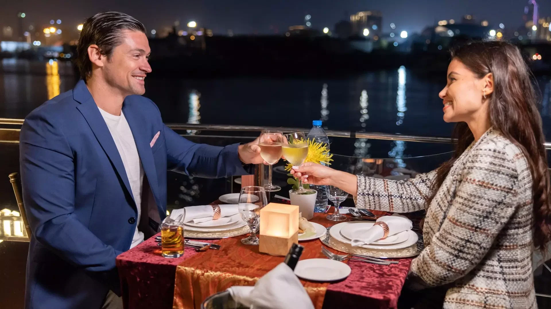 A well-dressed couple smiles and raises their wine glasses for a toast across a candlelit table at Dubai Water Canal Dhow Cruise.