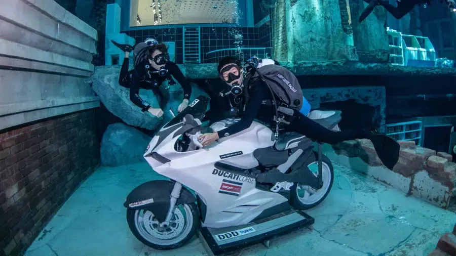 A scuba diver poses astride a submerged white motorcycle while another diver swims alongside underwater at Deep Dive Dubai.
