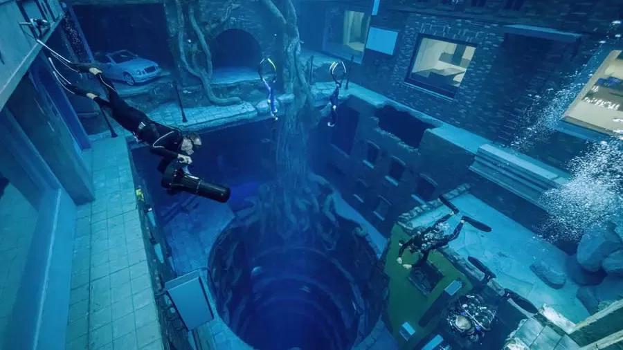 An elevated view shows multiple scuba divers swimming through an expansive sunken city structure with a large tree at Deep Dive Dubai.