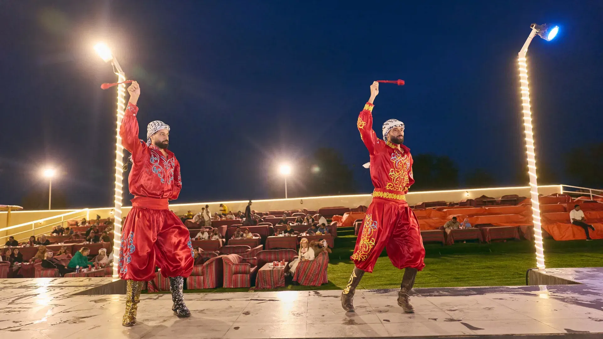 Two male performers in bright red tunics and matching pants hold up red props while standing on a stage at Desert Safari + Al Faqaa Camp.