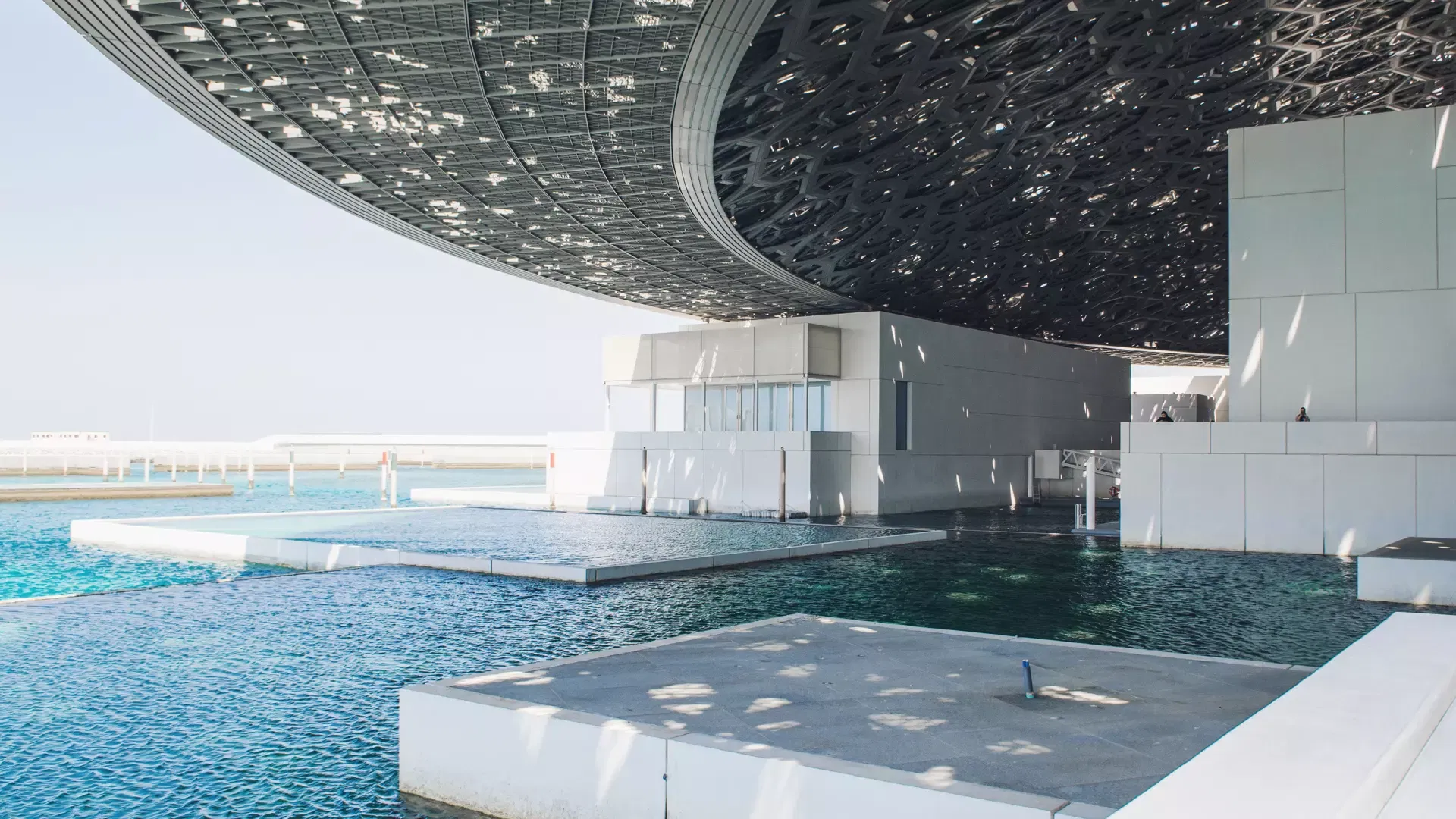 The curved underside of the dome overhanging turquoise seawater channels and low white gallery blocks at Louvre Abu Dhabi