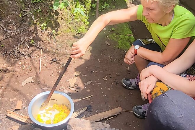 A visitor using a long wooden spoon to stir a pot of food cooking over a traditional stone fire in an outdoor setting.