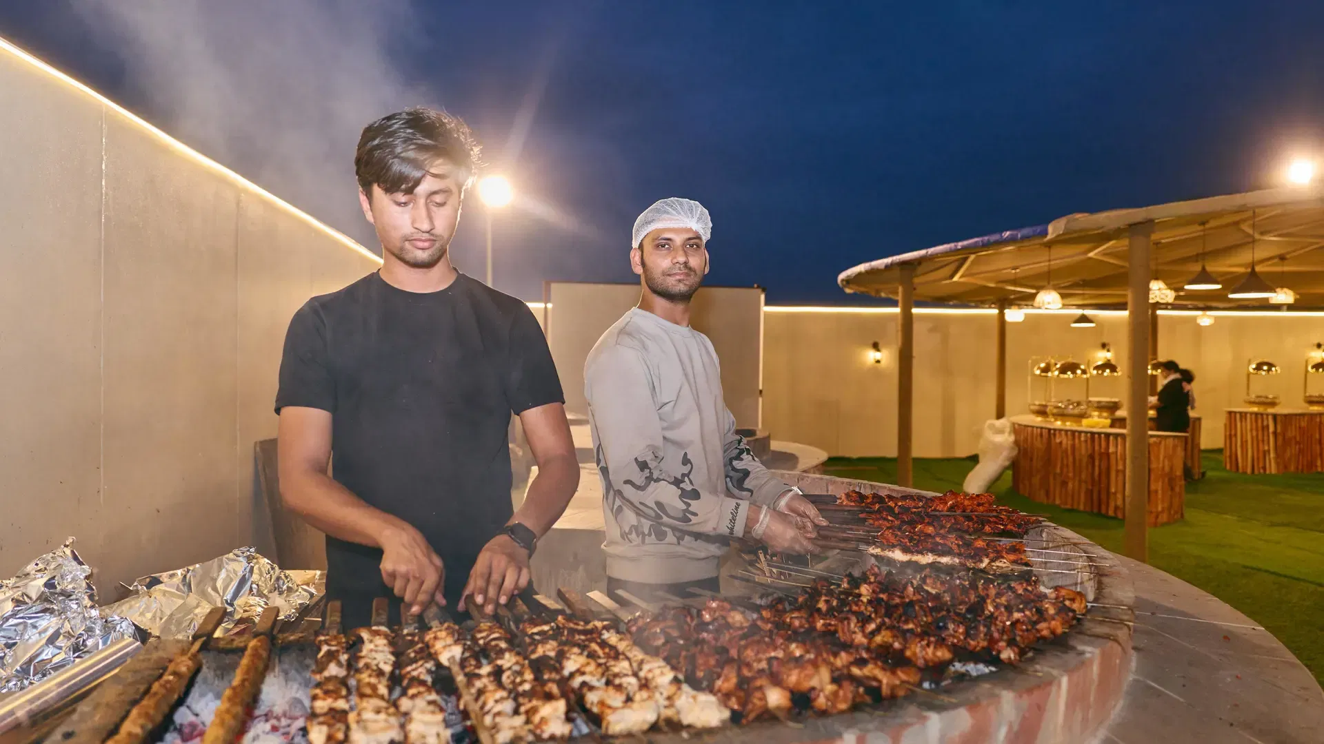 Two smiling chefs grill rows of meat skewers over glowing hot coals at Desert Safari + Al Faqaa Camp.