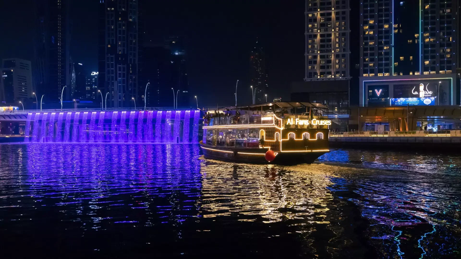 A brightly lit cruise boat sails past a waterfall cascading from a bridge illuminated in purple at Dubai Water Canal Dhow Cruise.